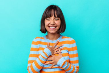 Mavi arka planda soyutlanmış genç melez bir kadın mutlu bir şekilde gülüyor ve ellerini karnının üstünde tutarak eğleniyor..