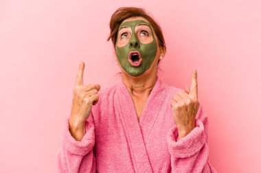 Middle age caucasian woman wearing a facial mask isolated on pink background pointing upside with opened mouth.