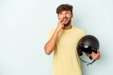 Mavi arka planda izole edilmiş motosiklet kaskı tutan genç bir ırk. Bir şeyin fotokopi alanına baktığını düşünerek rahatlamış..