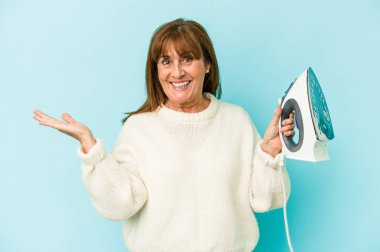 Middle age caucasian woman holding a iron isolated on blue background showing a copy space on a palm and holding another hand on waist.