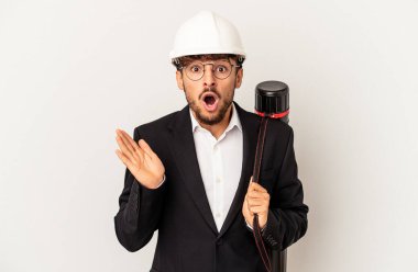 Gri arka planda izole edilmiş kask takan genç mimar şaşırmış ve şok olmuştu..