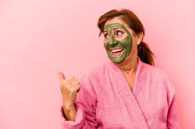 Middle age caucasian woman wearing a facial mask isolated on pink background points with thumb finger away, laughing and carefree.