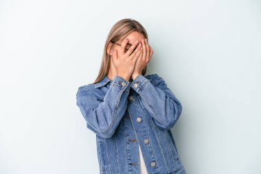 Mavi arka planda izole edilmiş genç beyaz kadın kameraya parmaklarıyla göz kırpıyor, yüzünü kapatmaktan utanıyor..