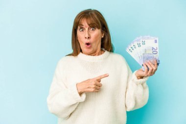 Middle age caucasian woman holding bank notes isolated on blue background pointing to the side