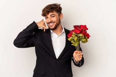 Beyaz arka planda izole edilmiş gül buketi tutan melez genç adam parmaklarıyla bir cep telefonu çağrısı hareketi gösteriyor..