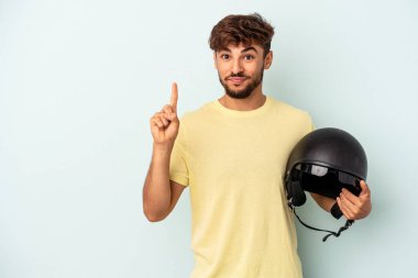 Mavi arka planda izole edilmiş motosiklet kaskı tutan genç melez bir numara parmağıyla görünüyor..