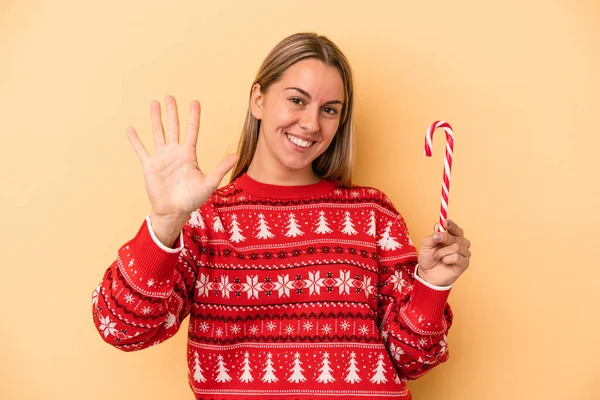 年轻的高加索女人拿着一根被黄色背景隔开的圣诞手杖 高兴地微笑着 用手指展示着5号 — 图库照片