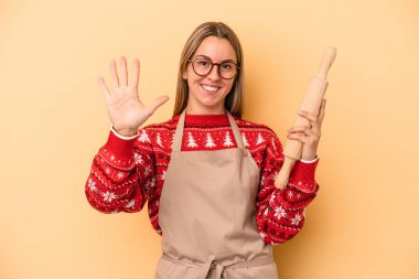 Genç fırıncı kadın, sarı arka planda izole edilmiş kurabiyeler yapıyor. Beş numarayı parmaklarıyla gülümsüyor..