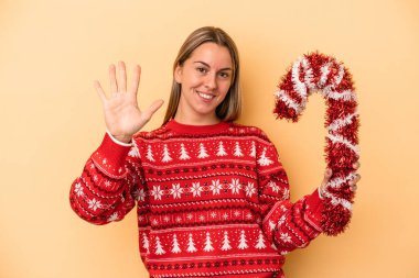 Sarı arka planda izole edilmiş büyük bir Noel sopası tutan beyaz bir kadın gülümseyerek beş numarayı parmaklarıyla gösteriyor..