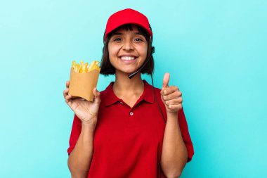 Genç melez kadın fast food restoranı çalışanı elinde kızartmalarla mavi arka planda izole edilmiş gülümsüyor ve başparmağını kaldırıyor.