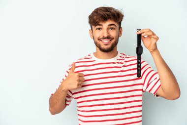 Mavi arka planda izole edilmiş bir kol saati tutan genç melez adam gülümsüyor ve başparmağını kaldırıyor.