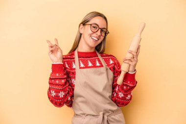 Genç fırıncı kadın Noel için kurabiye yapıyor sarı arka planda izole edilmiş neşeli ve kaygısız parmaklarıyla barış sembolü gösteriyor..