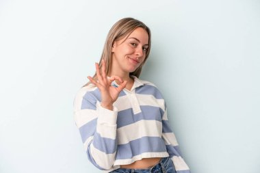 Kafkasyalı genç bir kadın mavi arka planda izole edilmiş bir şekilde bir göz kırpıyor ve elleriyle iyi bir jest yapıyor..