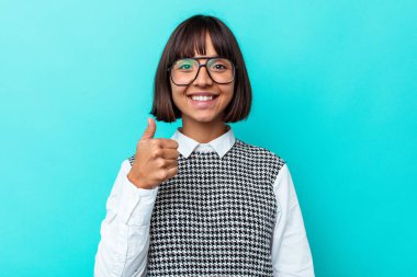 Mavi arka planda izole edilmiş genç melez kadın gülümsüyor ve başparmağını kaldırıyor.