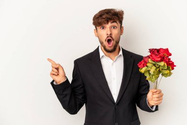 Beyaz arka planda izole edilmiş gül buketi tutan melez genç adam yan tarafı işaret ediyor.