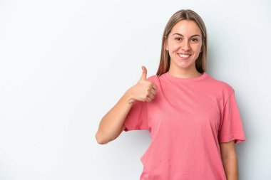 Mavi arka planda izole edilmiş genç beyaz kadın gülümsüyor ve başparmağını kaldırıyor.