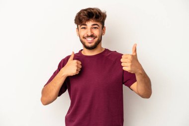 Genç melez adam gri arka planda izole edilmiş. Baş parmağını kaldırıyor, gülümsüyor ve kendinden emin..