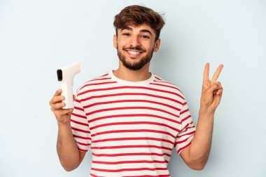 Mavi arka planda termometreyi tutan genç melez adam neşeli ve kaygısız parmaklarıyla barış sembolü gösteriyor..