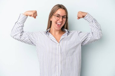 Mavi arka planda izole edilmiş genç beyaz kadın güçlü bir hareket sergiliyor kollarıyla, kadınsı gücün sembolü.