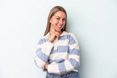 Mavi arka planda izole edilmiş genç beyaz kadın mutlu ve kendinden emin gülümsüyor, elleriyle çenesine dokunuyor..