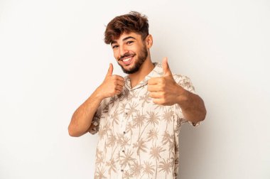 Genç melez adam gri arka planda izole edilmiş. Baş parmağını kaldırıyor, gülümsüyor ve kendinden emin..
