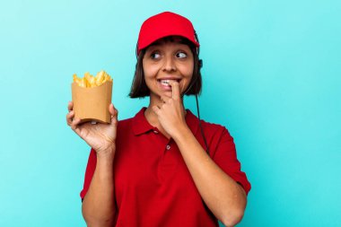 Genç melez kadın fast food restoranı çalışanı elinde kızartmalarla mavi arka planda izole edilmiş bir şekilde bir şeyin fotokopi odasına baktığını düşünerek rahatlıyor..
