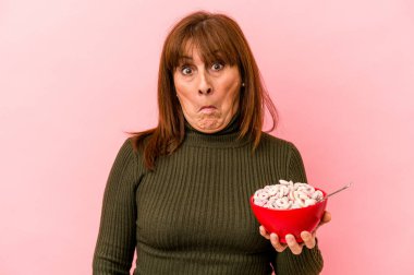 Middle age caucasian woman holding bowl of cereals isolated on pink background shrugs shoulders and open eyes confused.