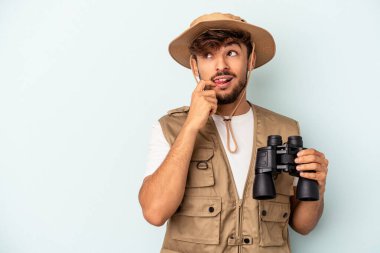 Young mixed race man holding binoculars isolated on blue background relaxed thinking about something looking at a copy space.
