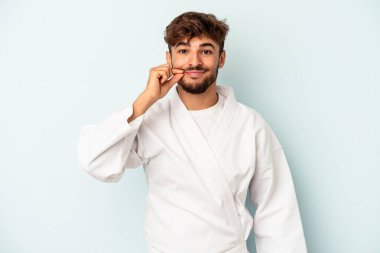 Young mixed race man doing karate isolated on blue background with fingers on lips keeping a secret.