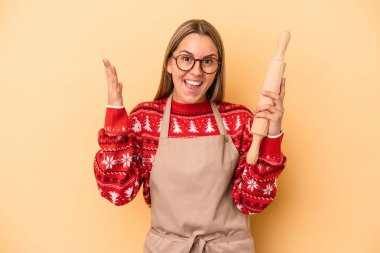 Genç fırıncı kadın, sarı arka planda izole edilmiş Noel için kurabiye yapıyor. Hoş bir sürpriz alıyor, heyecanlı ve el kaldırıyor..