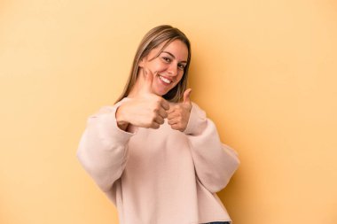 Sarı arka planda izole edilmiş genç beyaz kadın iki parmağını da kaldırıyor, gülümsüyor ve kendinden emin..