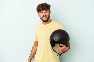 Young mixed race man holding motorcycle helmet isolated on blue background looks aside smiling, cheerful and pleasant.