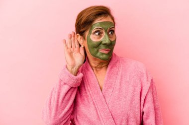 Middle age caucasian woman wearing a facial mask isolated on pink background trying to listening a gossip.