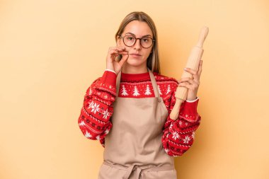 Genç fırıncı kadın Noel için kurabiye yapıyor sarı arka planda izole edilmiş dudaklarında parmaklarıyla bir sır saklıyor.