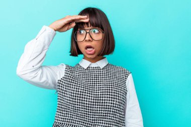 Genç melez kadın mavi arka planda izole edilmiş uzaklara bakıyor ve elini alnına koyuyor..