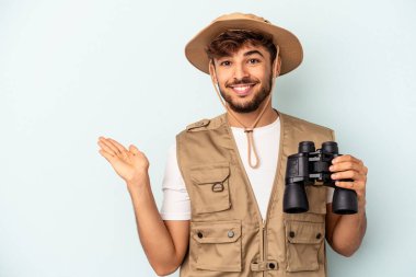 Young mixed race man holding binoculars isolated on blue background showing a copy space on a palm and holding another hand on waist.