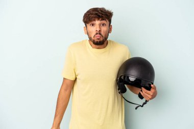 Mavi arka planda izole edilmiş motosiklet kaskı tutan genç bir ırk omuz silkiyor ve gözleri açık..