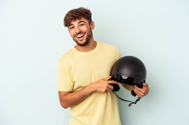 Mavi arka planda izole edilmiş motosiklet kaskı tutan genç melez adam gülüyor ve eğleniyor..