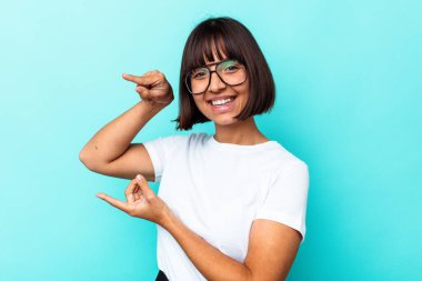 Mavi arka planda izole edilmiş genç melez bir kadın işaret parmaklarıyla küçük bir şey tutuyor, gülümsüyor ve kendinden emin..