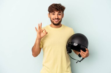 Mavi arka planda izole edilmiş motosiklet kaskı tutan genç melez adam neşeli ve kendinden emin bir şekilde iyi bir jest yapıyor..