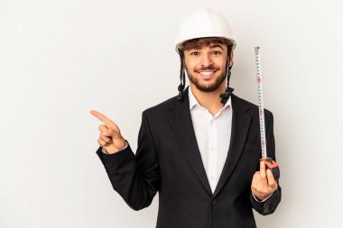 Miğfer takan ve taksimetre tutan genç mimar gri arka planda izole edilmiş gülümsüyor ve boş alanda bir şey gösteriyor..
