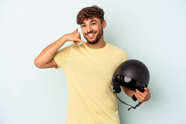 Mavi arka planda izole edilmiş motosiklet kaskı tutan melez genç bir adam parmaklarıyla bir cep telefonu hareketi gösteriyor..