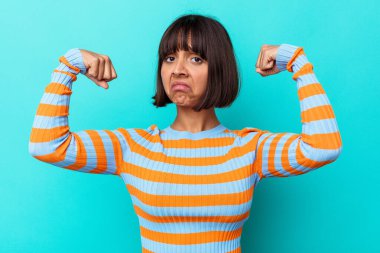 Mavi arka planda izole edilmiş genç bir ırk kadını güçlü bir jest yapıyor, kadınsı gücün sembolü.