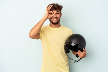 Mavi arka planda izole edilmiş motosiklet kaskı tutan genç melez adam heyecanlı..