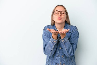 Mavi arka planda izole edilmiş genç, beyaz bir kadın. Dudaklarını kıvırıyor ve avuç içlerine hava öpücüğü yolluyor..