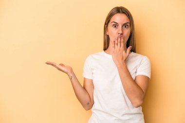 Sarı arka planda izole edilmiş genç beyaz kadın avuç içinin fotokopi alanını elinde tutmaktan etkilenmiş..