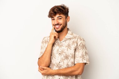 Genç melez adam gri arka planda izole edilmiş mutlu ve kendinden emin gülümsüyor, elleriyle çenesine dokunuyor..