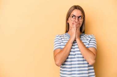 Genç beyaz kadın, sarı arka planda izole edilmiş. Düşünceli bir şekilde, ağzı elle kapatılmış bir fotokopi odasına bakıyor..