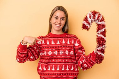 Elinde büyük bir noel sopası tutan beyaz bir kadın sarı arka planda izole edilmiş, gömlek fotokopi odasını işaret eden gururlu ve kendinden emin bir kadın.