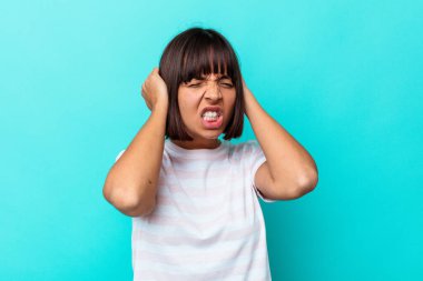Mavi arka planda izole edilmiş genç bir ırk kadını kulaklarını elleriyle kapatıyor..